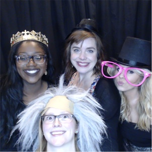 Four women in a photo booth at a party. One wearing a fun hat, one wearing the same hat and huge pink glasses, another one wearing a crown, and one wearing a crazy scientist wig.